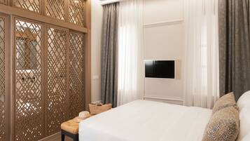 Una habitación de hotel con una cama, un televisor montado en la pared, un separador de madera y un banco con una cesta.