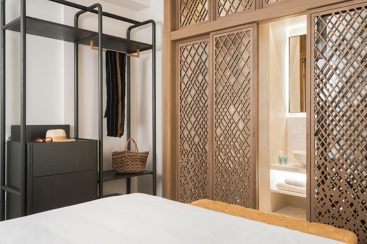 A bedroom with a bed, a wardrobe, a shelf with a hat and a basket, and a bathroom visible through a wooden lattice partition.