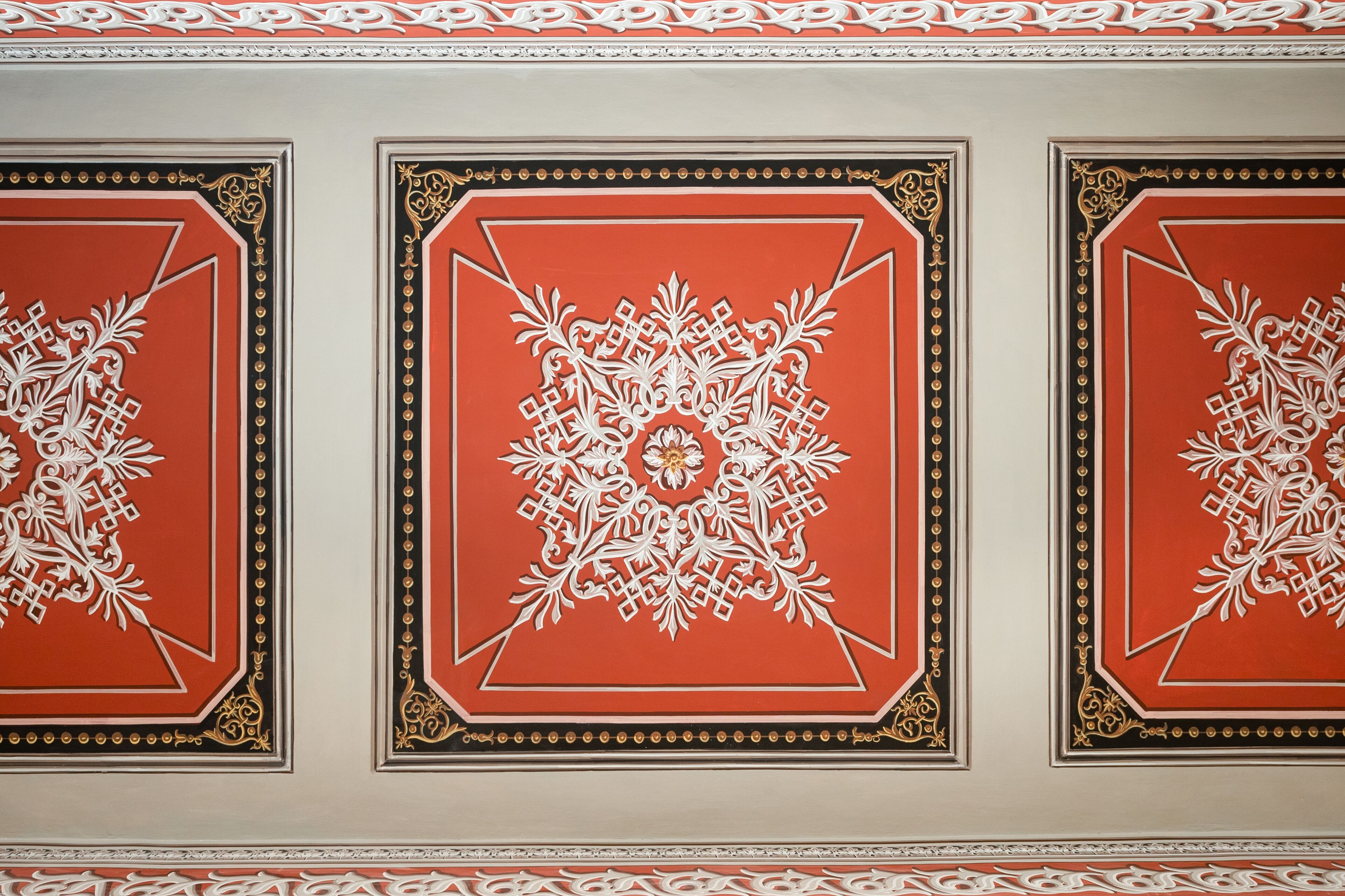 Three decorative panels with intricate white patterns on a red background, framed by black and gold borders.