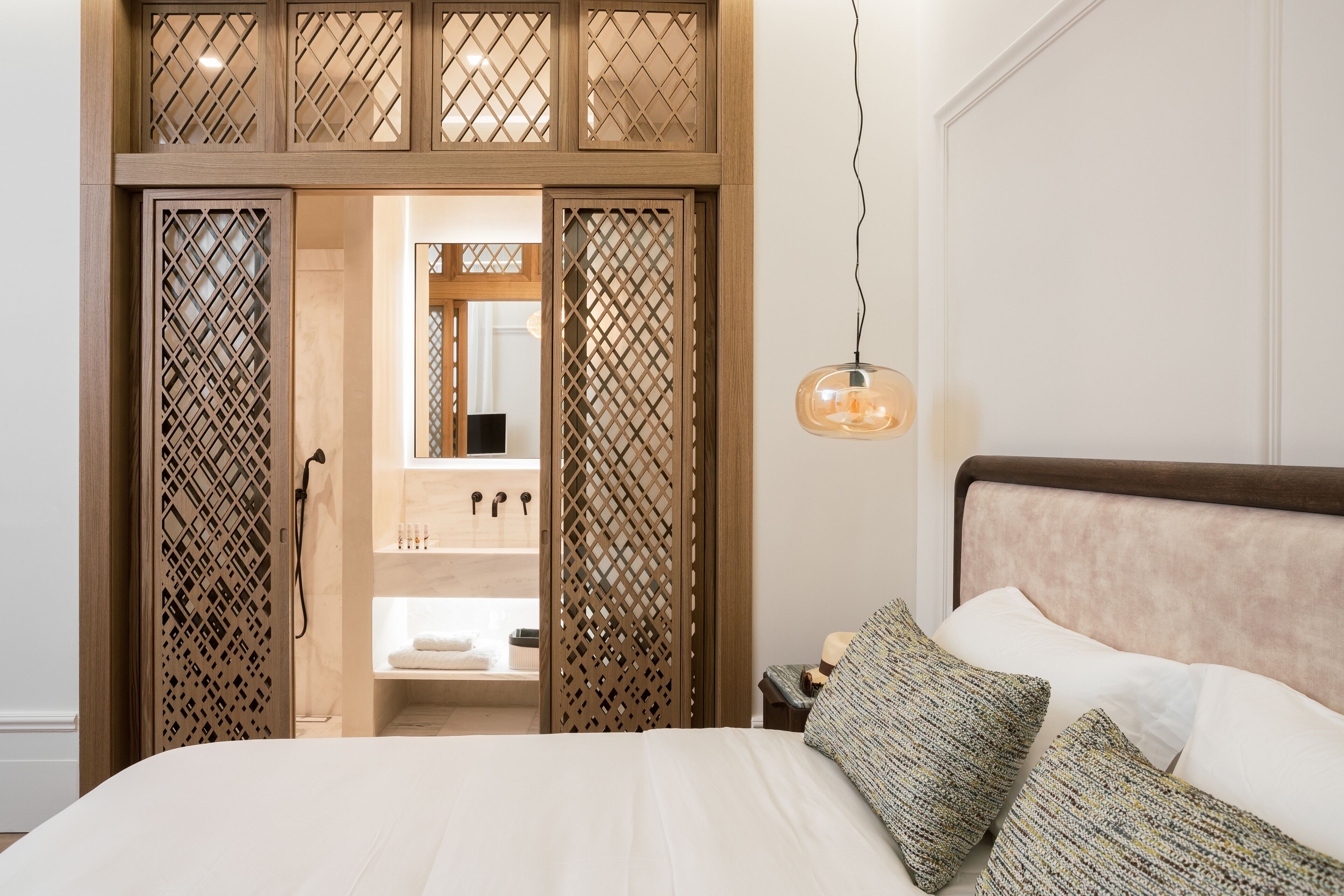 A bedroom with a bed, pillows, a hanging light fixture, and a wooden partition with a bathroom visible through it.