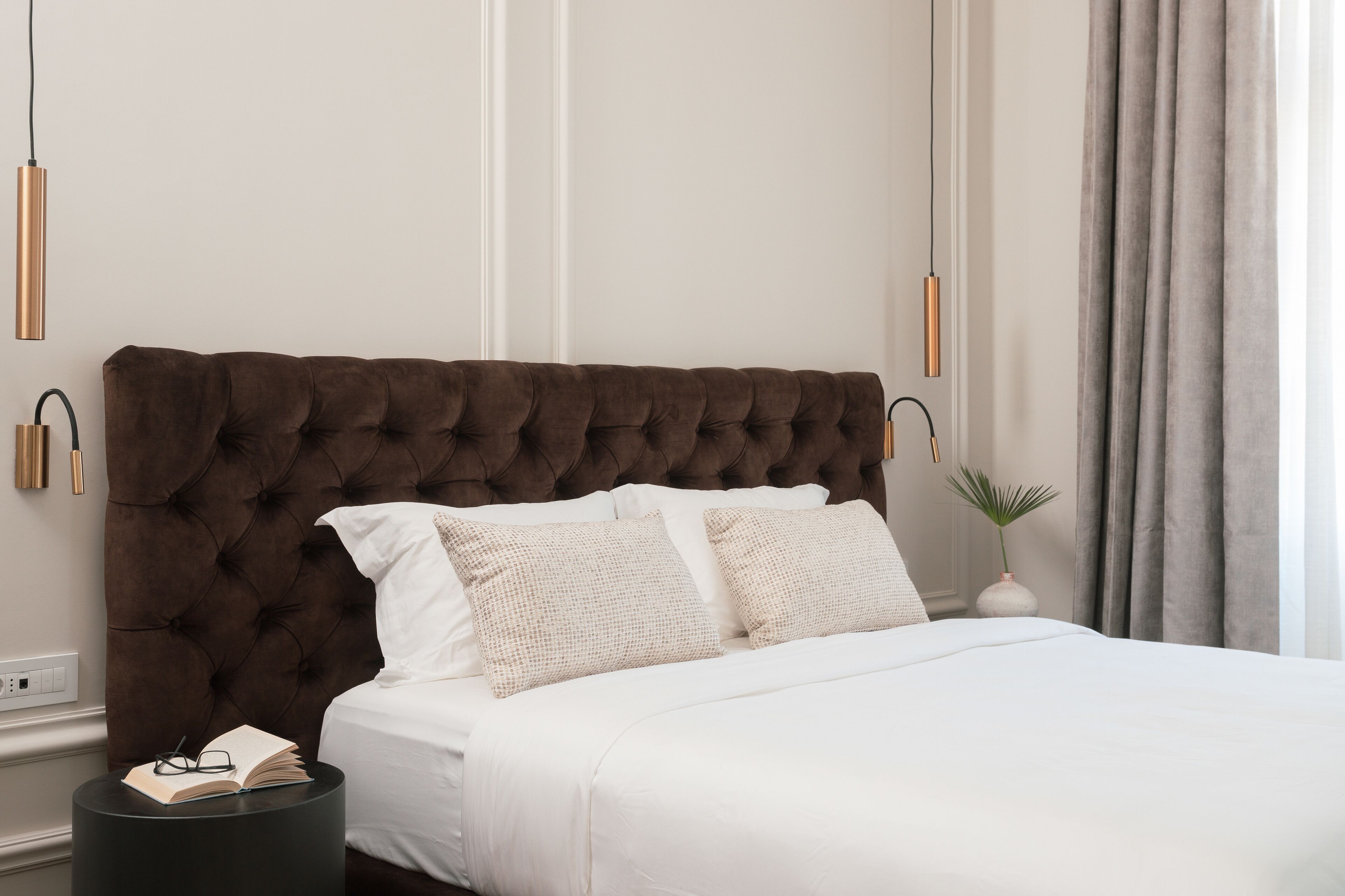 A bedroom with a tufted headboard, white bedding, a bedside table with a book and glasses, and a plant.