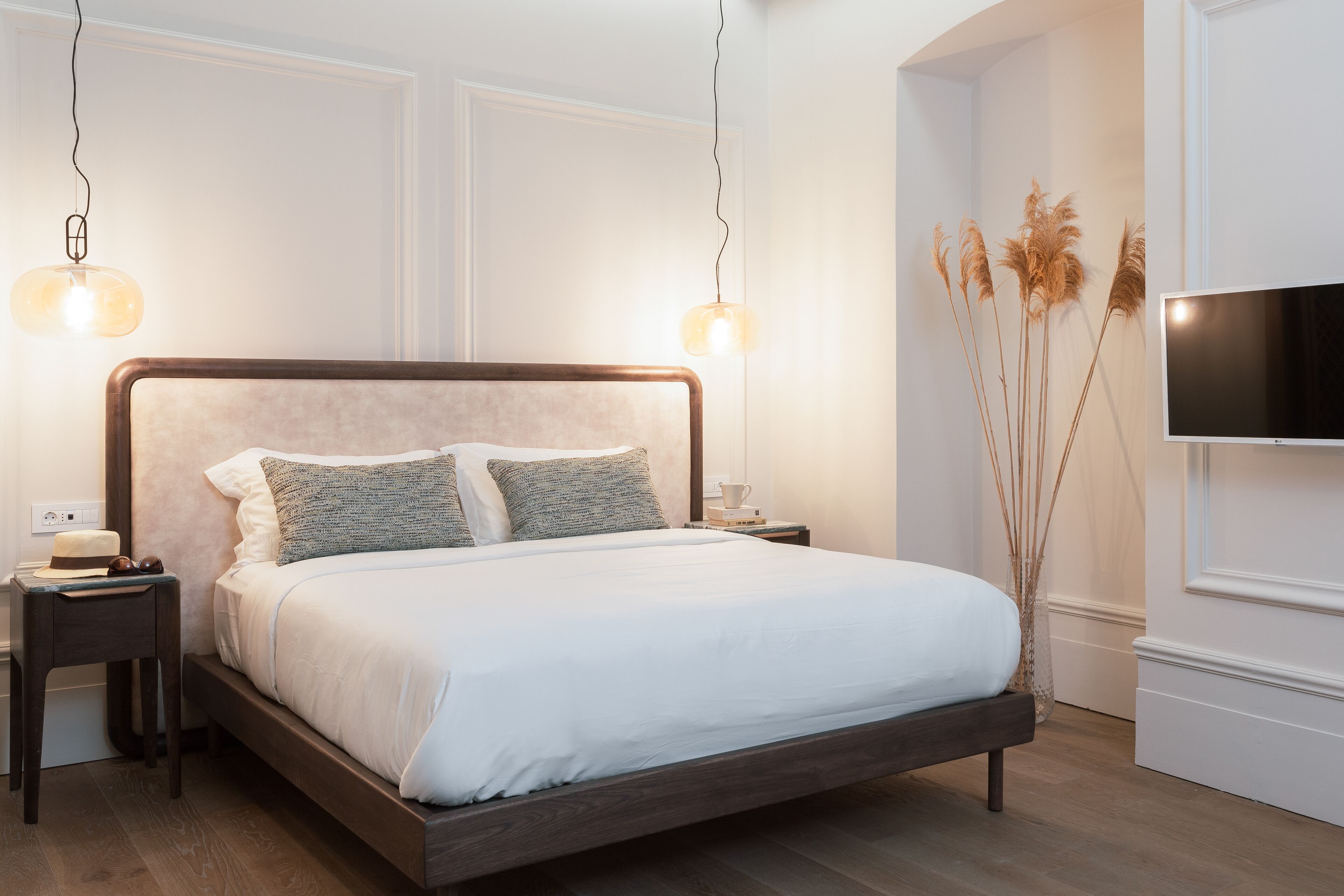 A neatly made bed with a wooden frame and white bedding, two bedside tables with lamps, and a flat-screen TV mounted on the wall.
