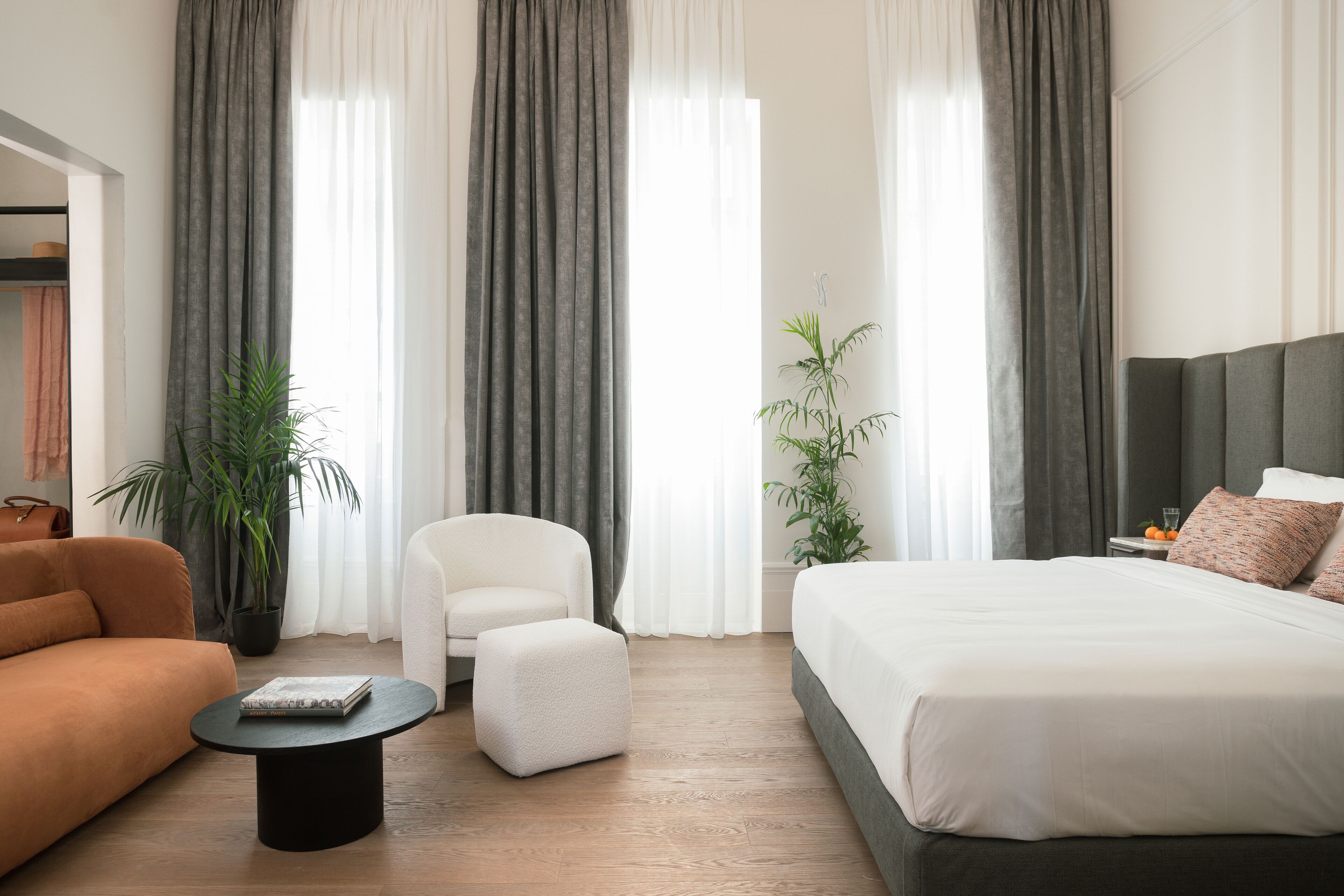 A modern hotel room with a bed, a sofa, a chair, a small table, and two potted plants.