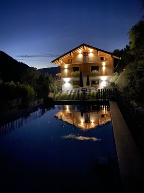 Una casa de madera con balcón, rodeada por una valla, y su reflejo en una piscina.