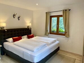Una cama bien hecha con sábanas blancas y almohadas rojas, un cabecero capitonado, dos lámparas de pared y una ventana con cortinas.