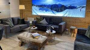 A modern living room with a large flat-screen TV displaying a snowy mountain scene, a grey sofa, a round coffee table, and a tufted ottoman. - ALM SEASONS - Family Chalet-Studio with sauna (Saalbach)