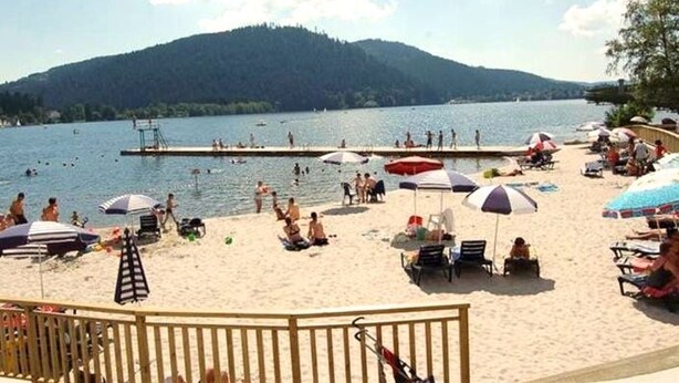 A beach with people swimming and sunbathing, a wooden deck, and a mountain in the background. - Cozy chalet in Gérardmer, ski view, 4 guests (Gérardmer)
