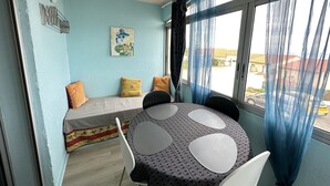 Ein Zimmer mit Esstisch, Stühlen, einer Couch mit Kissen, einem Fenster mit Vorhängen und einer Wanduhr.