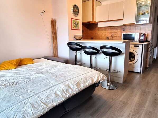 A compact hotel room with a bed, a small kitchenette, a washing machine, and a bar area with stools.