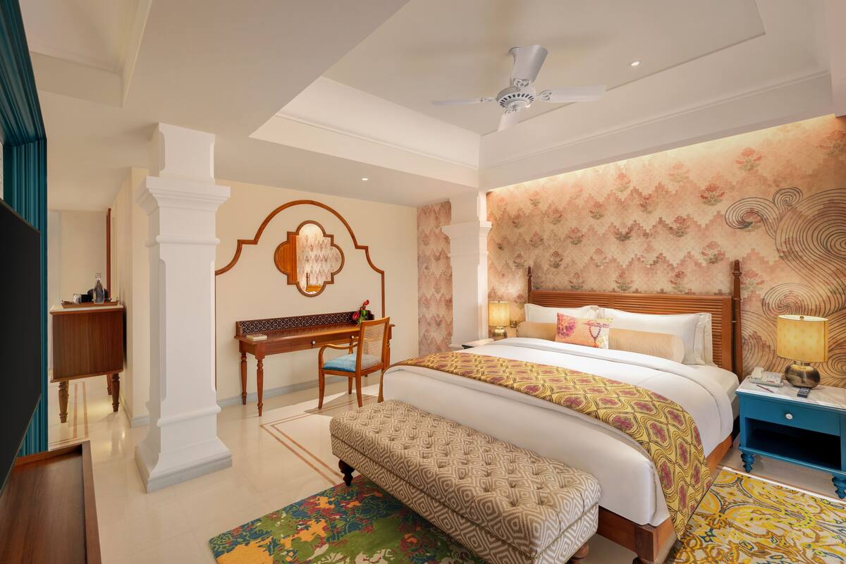 A hotel room with a large bed, a desk, a chair, a bench, a nightstand, and a ceiling fan.