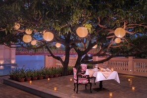 An outdoor dining area with a table set under a tree, surrounded by potted plants and lit by candles.
