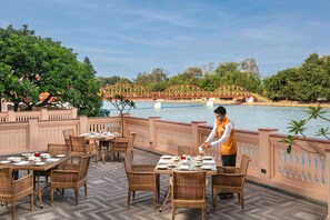 A dining area with tables set for guests, overlooking a river with a bridge, surrounded by lush greenery.