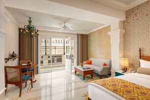 A hotel room with a bed, a sofa, a dining table with chairs, a chandelier, a ceiling fan, and a view of the outside balcony.