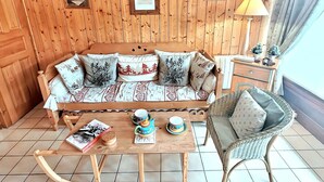 Et hyggeligt rum med en træpanelvæg, en sofa med mønstrede puder, en fletstol, et lille bord med te og kopper, og en lampe på et sengebord.