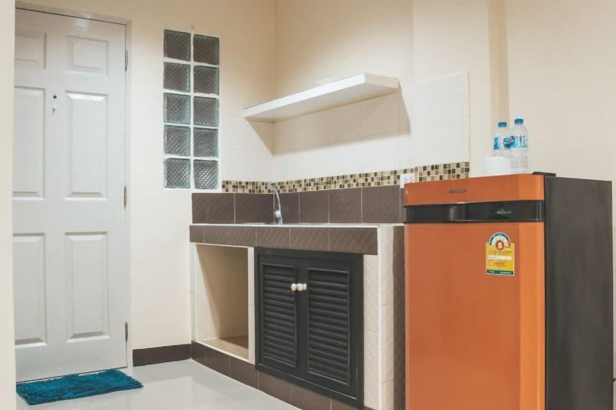 A compact kitchen with a refrigerator, a microwave, and a small sink. There is a door with a window, a shelf above the sink, and a blue rug on the floor.