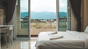 A bedroom with a bed, a desk, and a chair. There is a balcony with a view of mountains and houses. - Nattida Place (Chiang Mai)