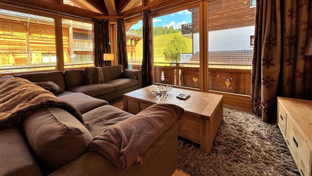 A cozy log cabin interior with a sofa, a wooden coffee table, and large windows offering a view of a green landscape. - Spacious apartment in village center with parking (Les Gets)