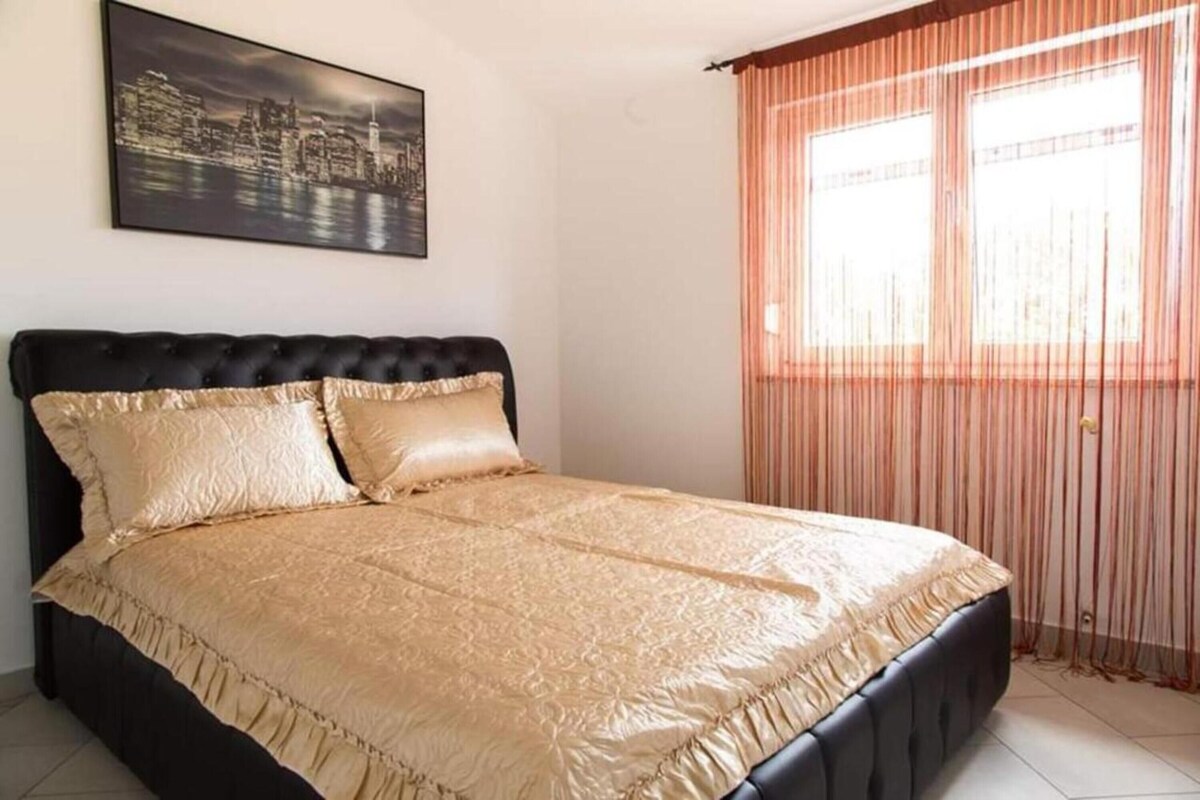 A bedroom with a large bed, a cityscape painting on the wall, and a window with curtains.