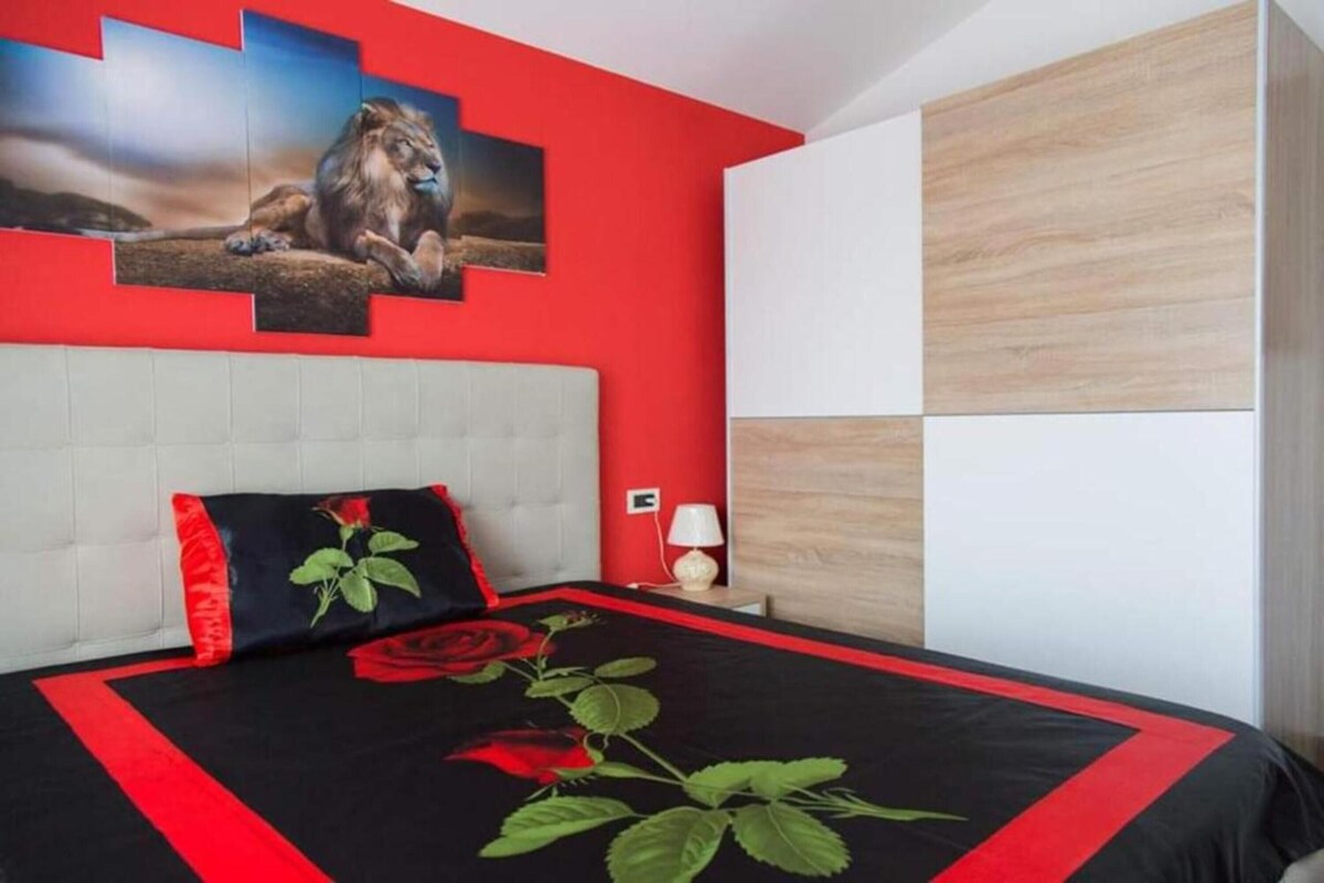 A bedroom with a red accent wall, a white headboard, a bed with a black and red rose-themed bedspread, and a nightstand with a lamp.