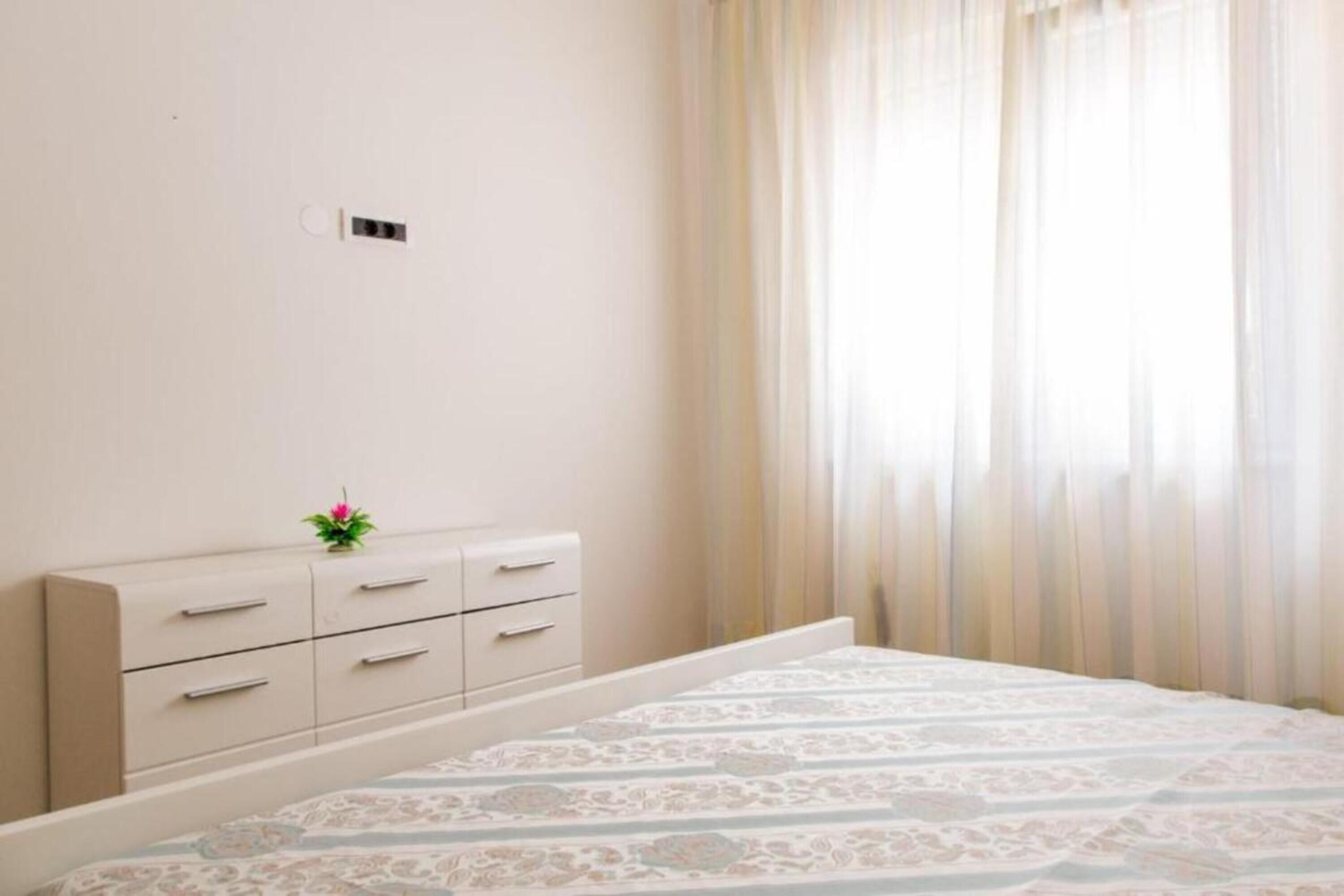 Un dormitorio con cama, mesita de noche, una planta pequeña y una ventana con cortinas de encaje.