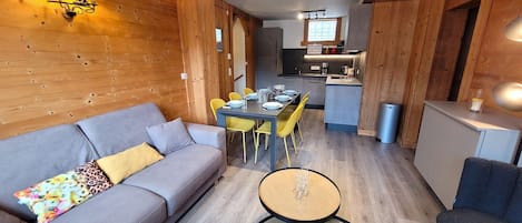 A modern living room with a grey sofa, a round coffee table with two bowls, a dining area with yellow chairs, and a kitchen in the background.
