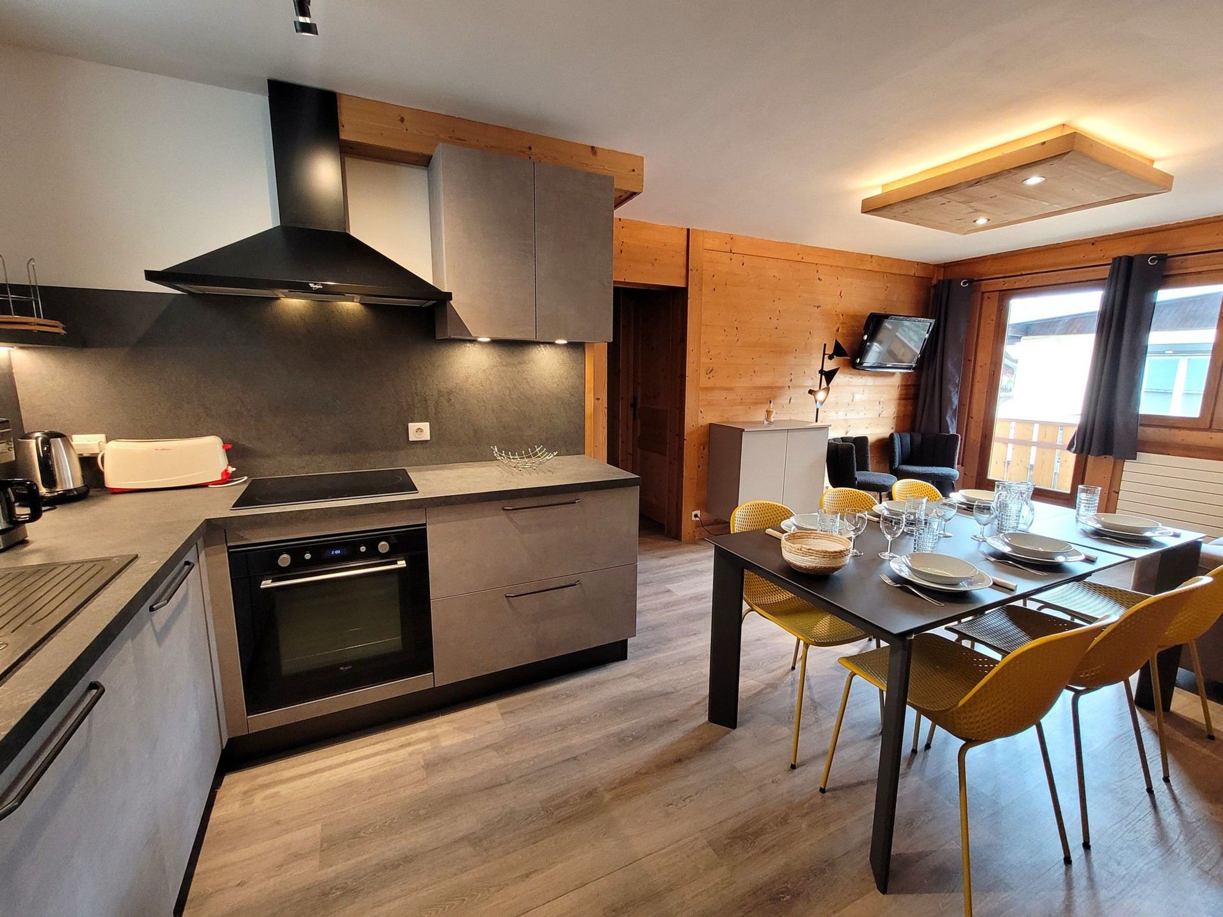 A modern kitchen with a central island, built-in oven, and stainless steel appliances. There is a dining area with yellow chairs and a table set for four.