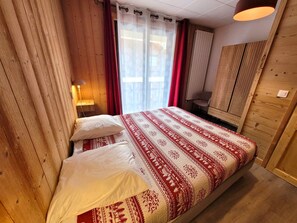 Un dormitorio con suelo de madera, una cama con colcha estampada, una lámpara de noche, una ventana con cortinas y un radiador.