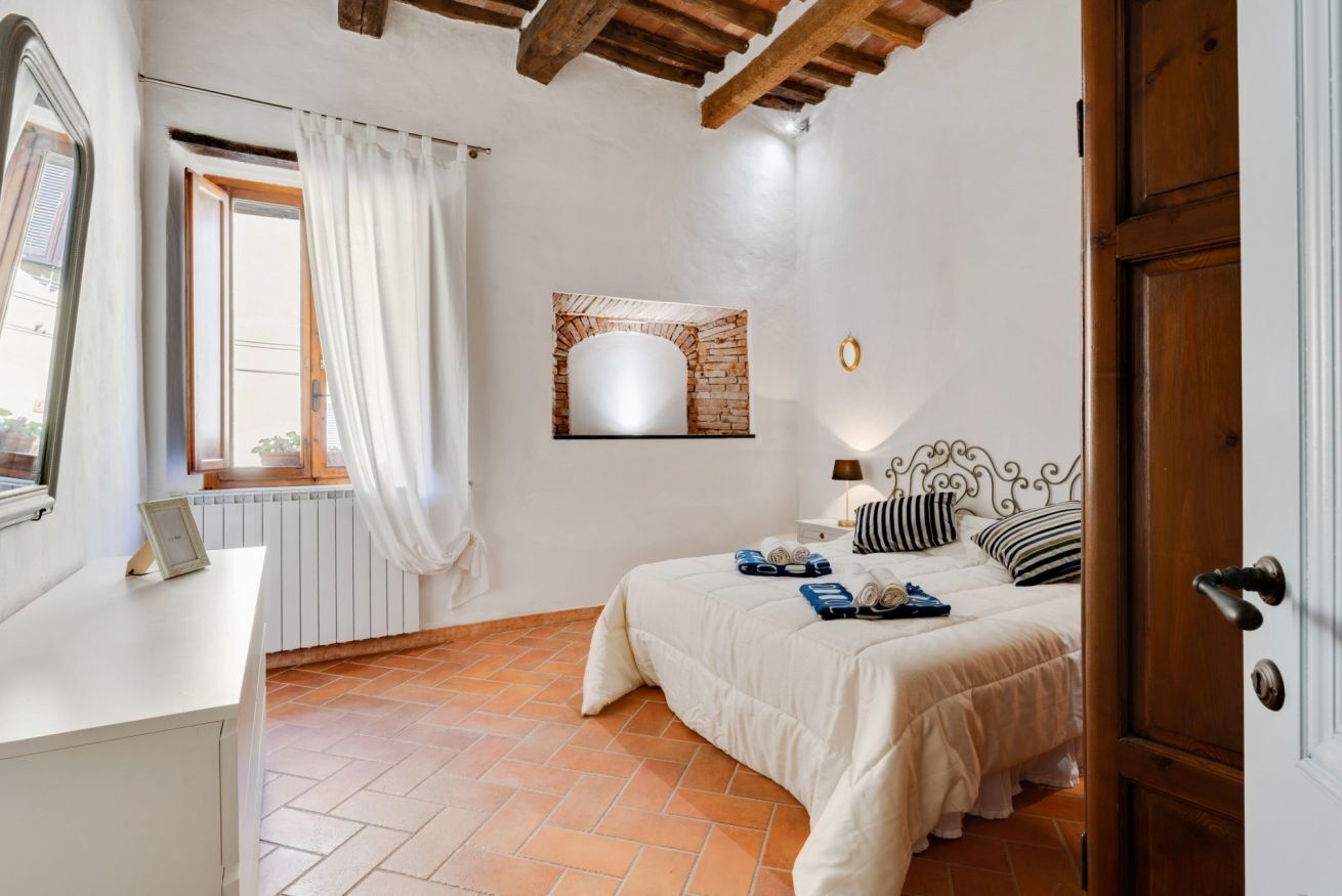 Quarto com cama, janela com cortinas, radiador, espelho fixado na parede e porta.
