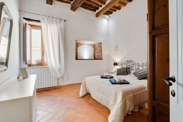 A bedroom with a bed, a window with curtains, a radiator, a wall-mounted mirror, and a door.