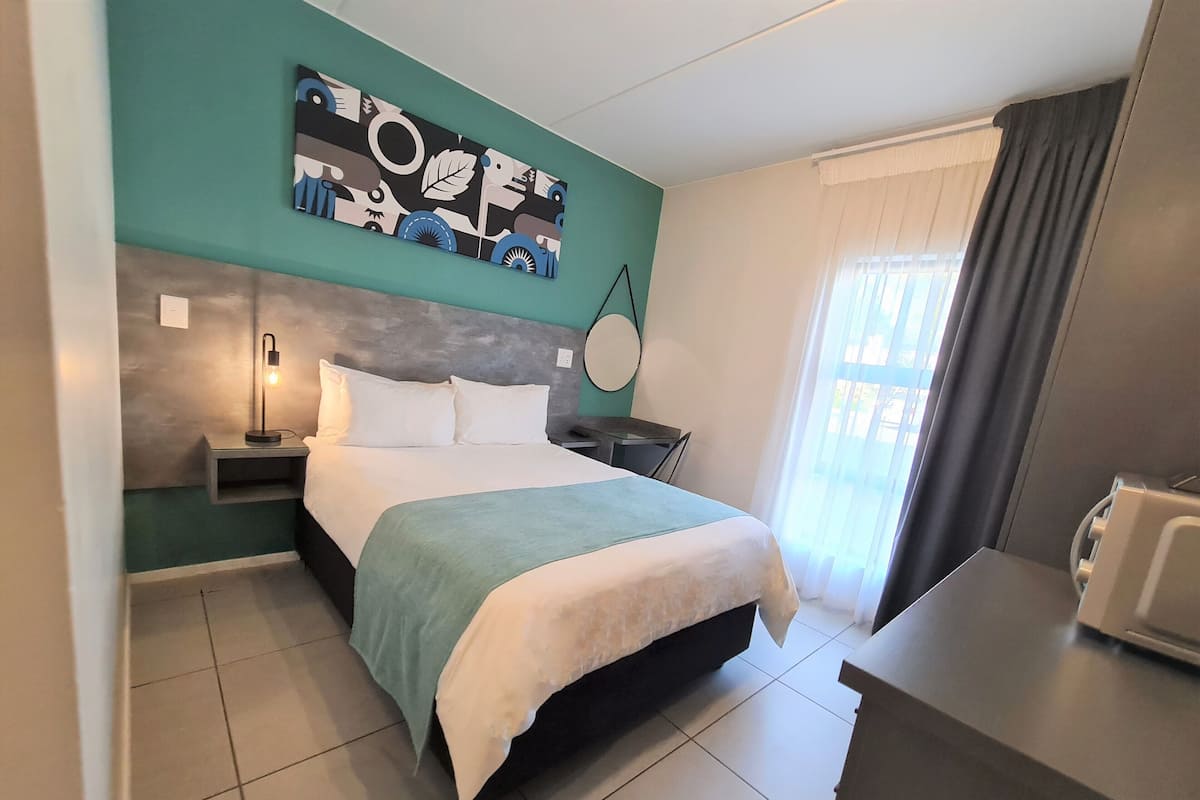A modern hotel room with a large bed, a bedside table with a lamp, a desk with a chair, and a window with curtains.
