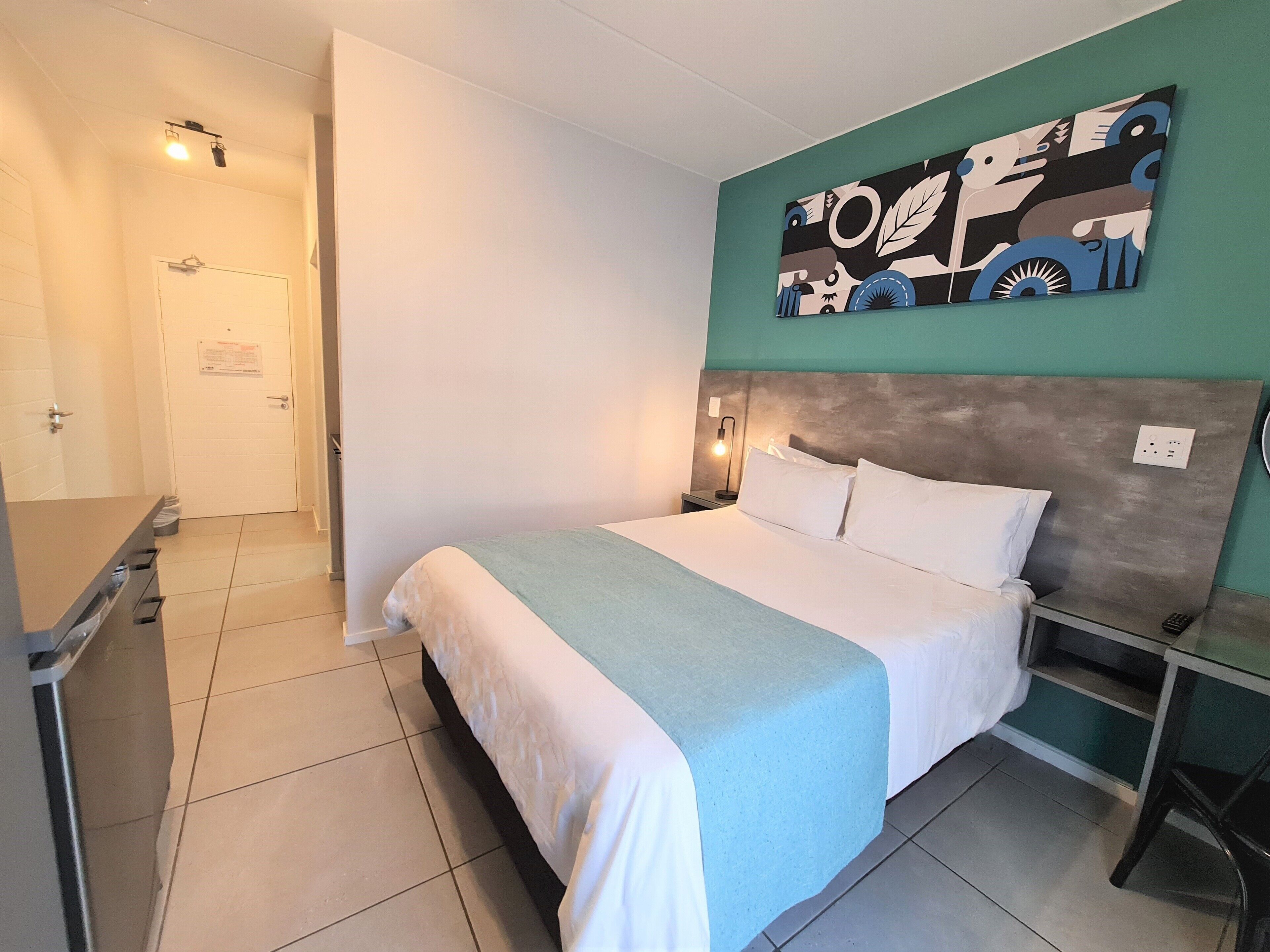 A hotel room with a bed, a desk, a chair, a nightstand, and a wall art piece.