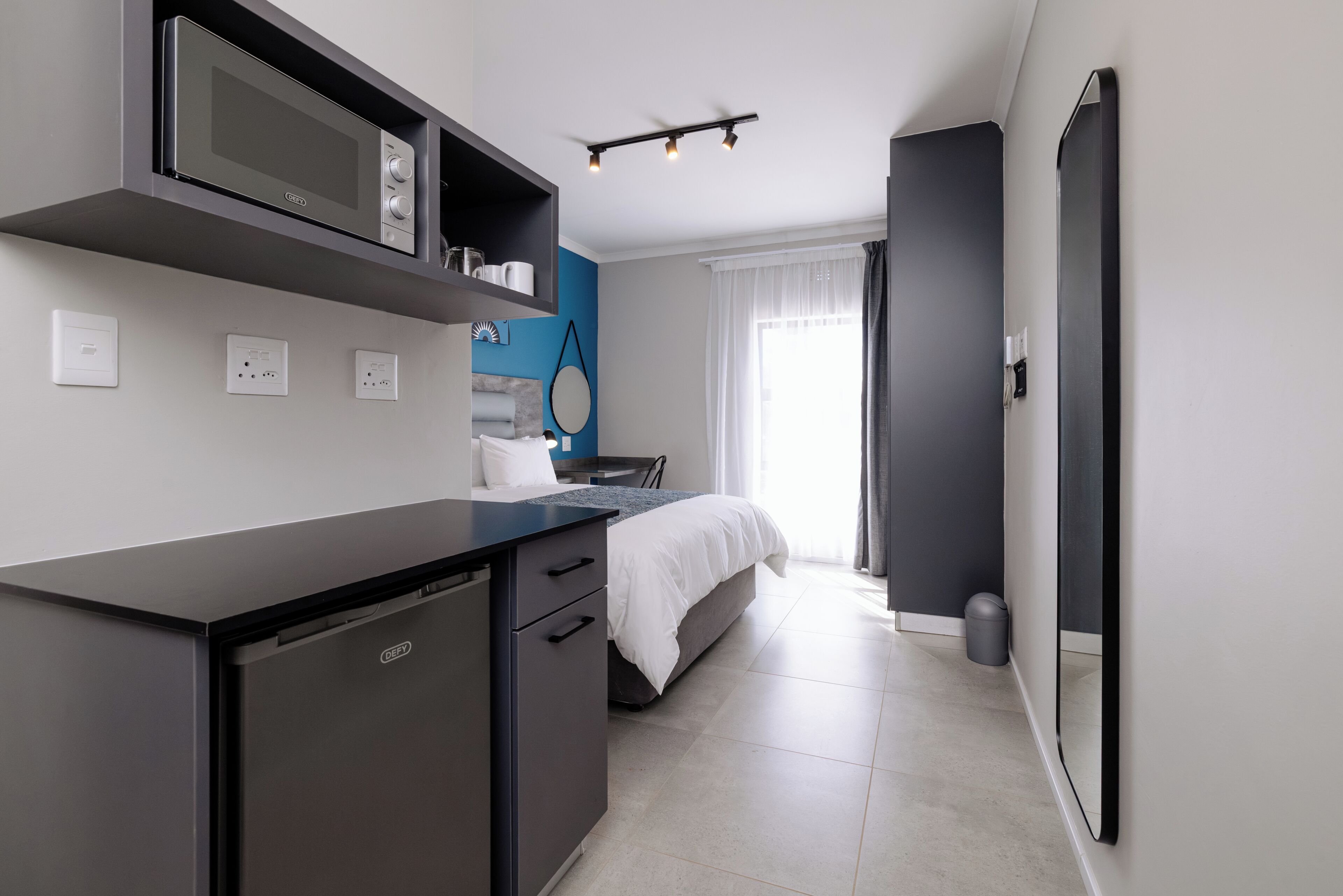 Una cocina moderna con microondas, horno empotrado y cafetera. Hay una cama con cubrecama blanco y una mesita de noche pequeña. La habitación tiene un ventanal con cortinas, un espejo y un aplique de pared.