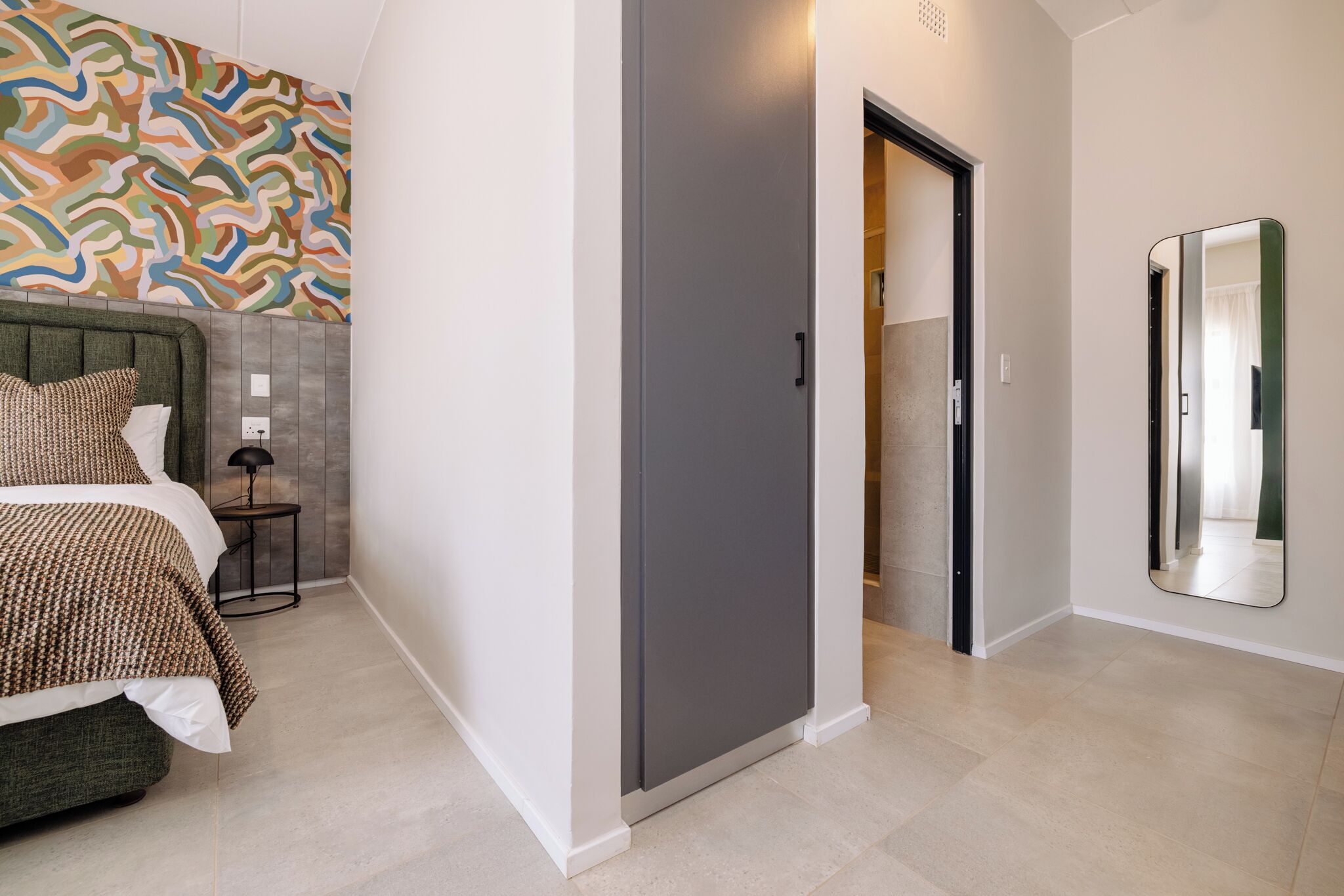 A bedroom with a bed, a bedside table, a wall with a colorful abstract pattern, a grey wardrobe, and a mirror.