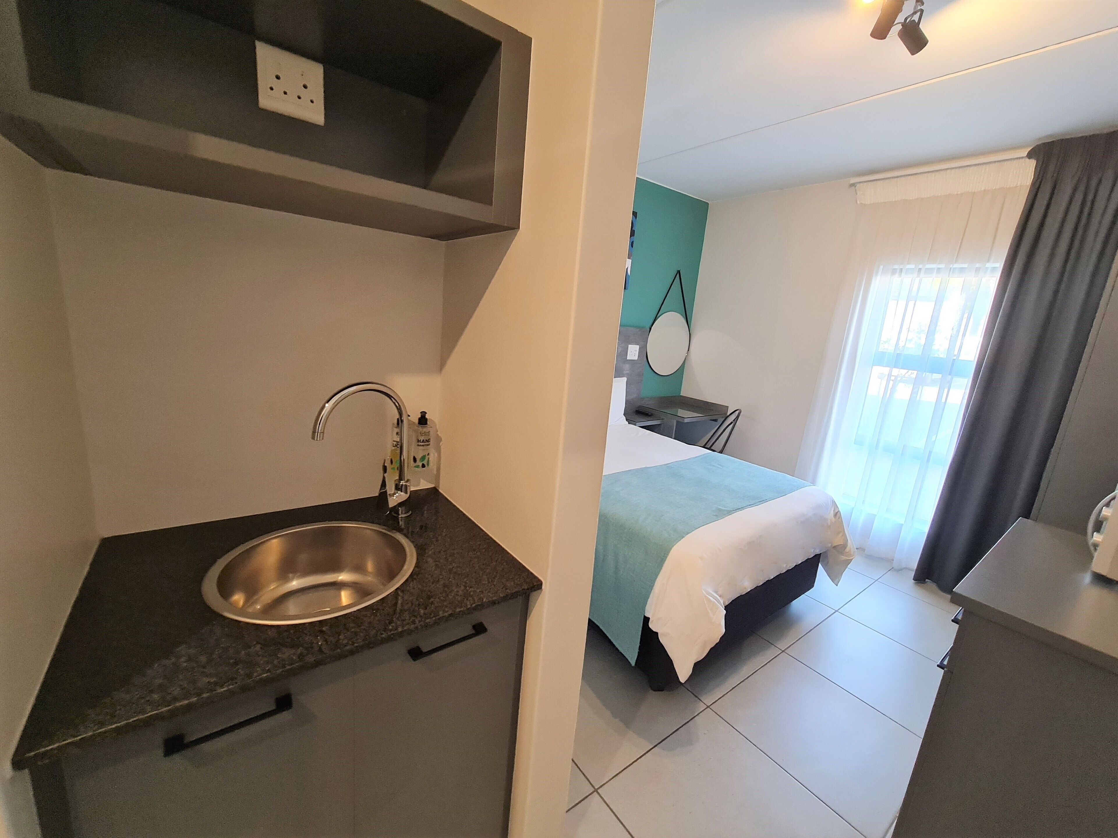 A modern hotel room with a kitchenette, a bed, a bedside table, and a window with curtains.