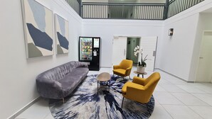 Una sala de estar moderna con un sofá gris, dos sillones amarillos, una mesa de centro y una alfombra. Se ve una máquina expendedora al fondo.