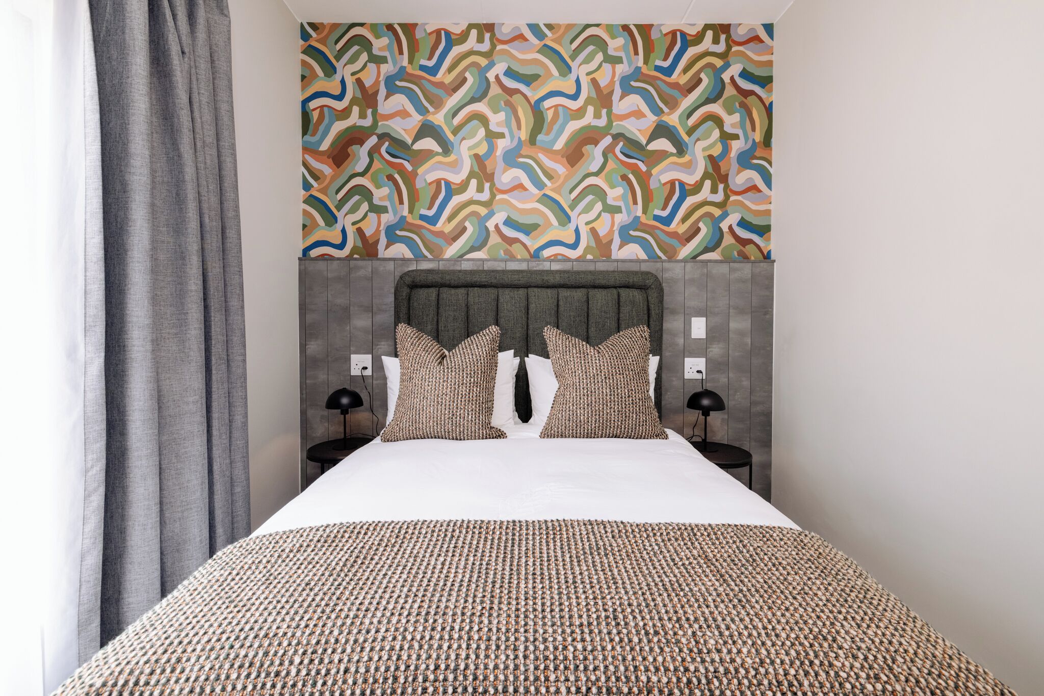 Una habitación de hotel moderna con una cama grande, un cabecero estampado y un diseño abstracto y colorido en la pared.