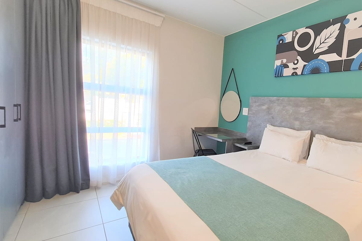 A hotel room with a bed, a desk, a chair, a window with curtains, and a wall with a circular mirror.