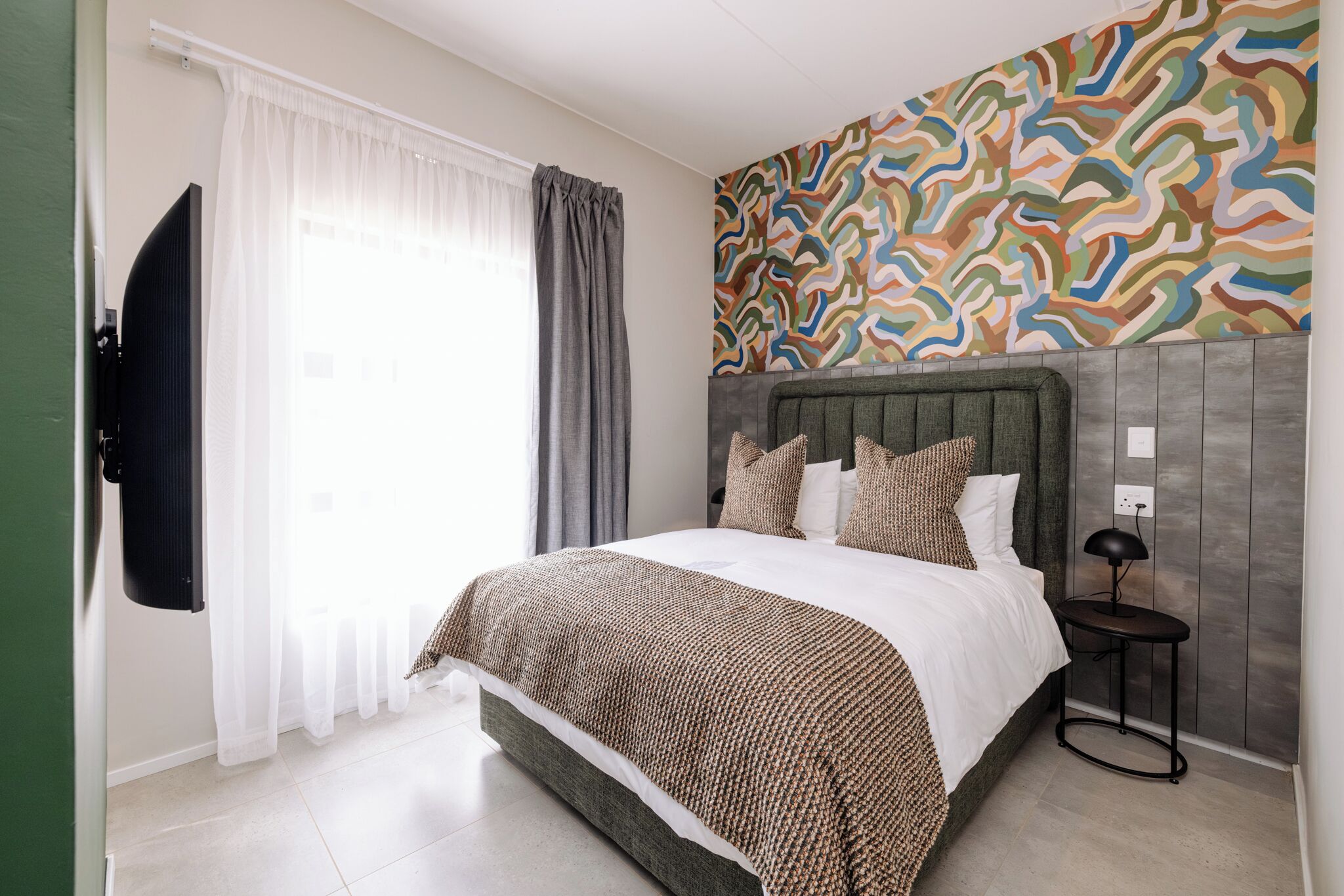 A bedroom with a bed, a television, a bedside table, and a wall with a colorful abstract pattern.