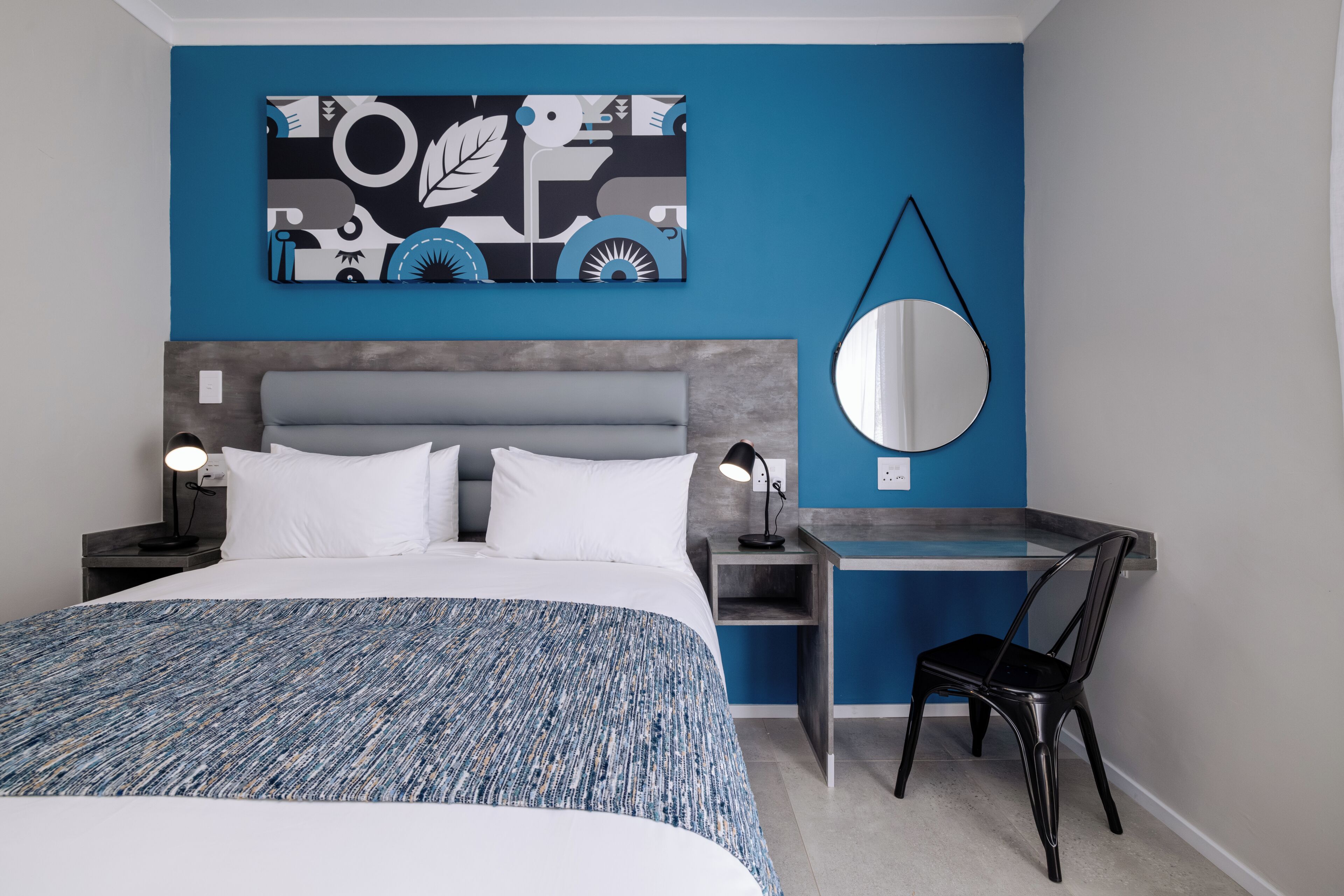 Una habitación de hotel moderna con una cama, un escritorio, una silla, un espejo redondo y una obra de arte en la pared.