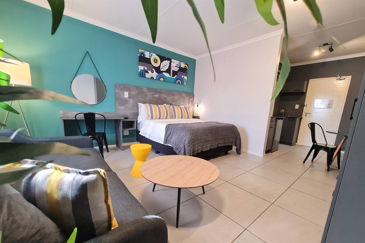 A modern hotel room with a bed, a desk, a chair, a round table, and a kitchenette.