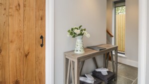 Une porte en bois, un vase blanc rempli de fleurs, une petite table et une fenêtre donnant sur des arbres.