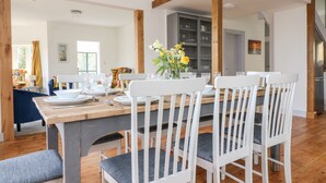 Comedor con mesa de madera dispuesta para cuatro, sillas blancas con cojines grises y un jarrón con flores amarillas.