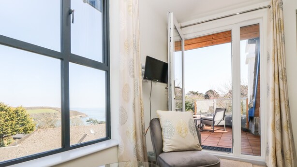 Una habitación con una mesa de vidrio, una silla gris con cojín, un televisor en la pared y vistas al mar a través de una puerta corrediza de vidrio.