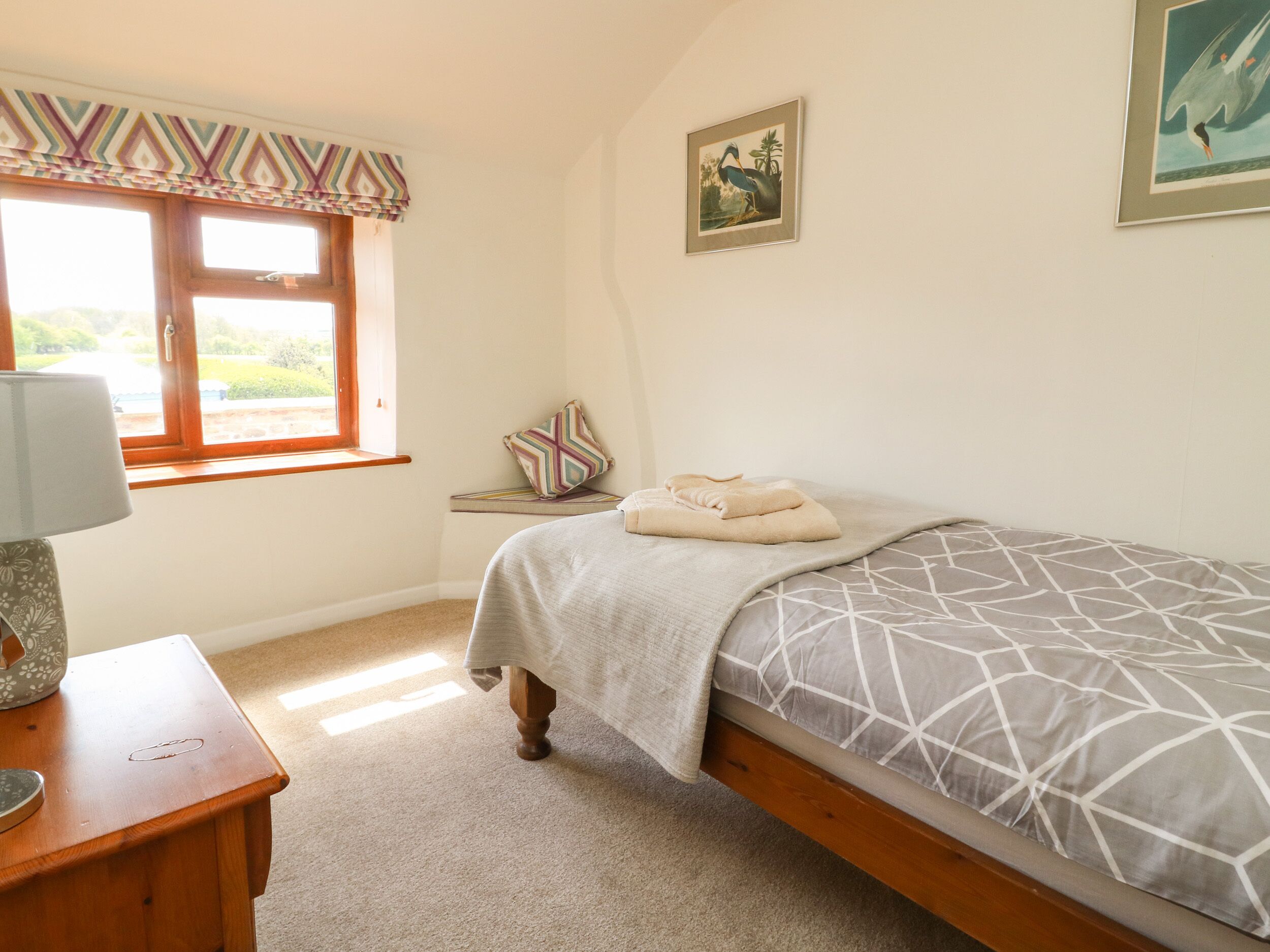 Un dormitorio con una cama, una mesita de noche, una lámpara, una ventana con cortinas, un cuadro en la pared y un pequeño estante con un almohadón.