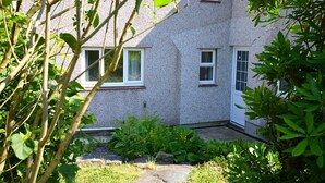 A two-story building with a textured exterior, multiple windows, and a garden area with various plants and a stone pathway. - Idan House (Llanfairpwllgwyngyll)