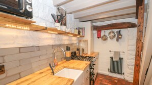 Une cuisine avec un plan de travail en bois, un évier, une cuisinière et un sol en briques. Il y a des étagères au-dessus du plan de travail et un radiateur au mur.