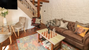 Un salon chaleureux avec un canapé marron, une table basse en verre, une table à carreaux et un tapis à motifs. On aperçoit, au fond, un escalier avec des poutres en bois et un mur blanc.