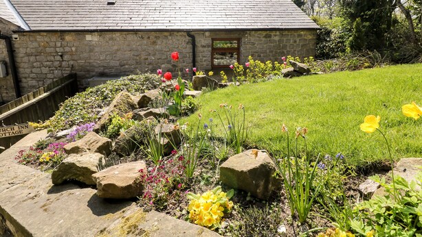 Una cabaña de piedra con techo inclinado, rodeada de un jardín cuidado con diversas flores y rocas.