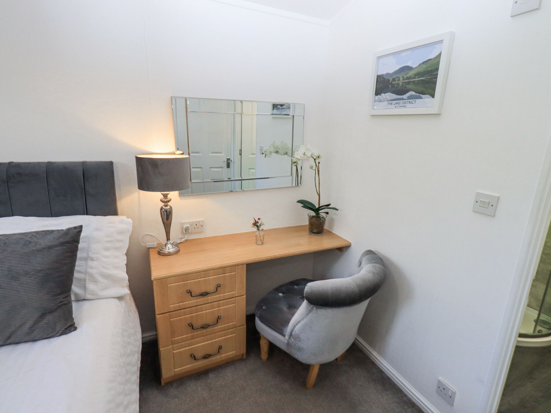 A bedroom with a bed, a bedside lamp, a wooden desk with drawers, a chair, a mirror, and a framed picture on the wall.