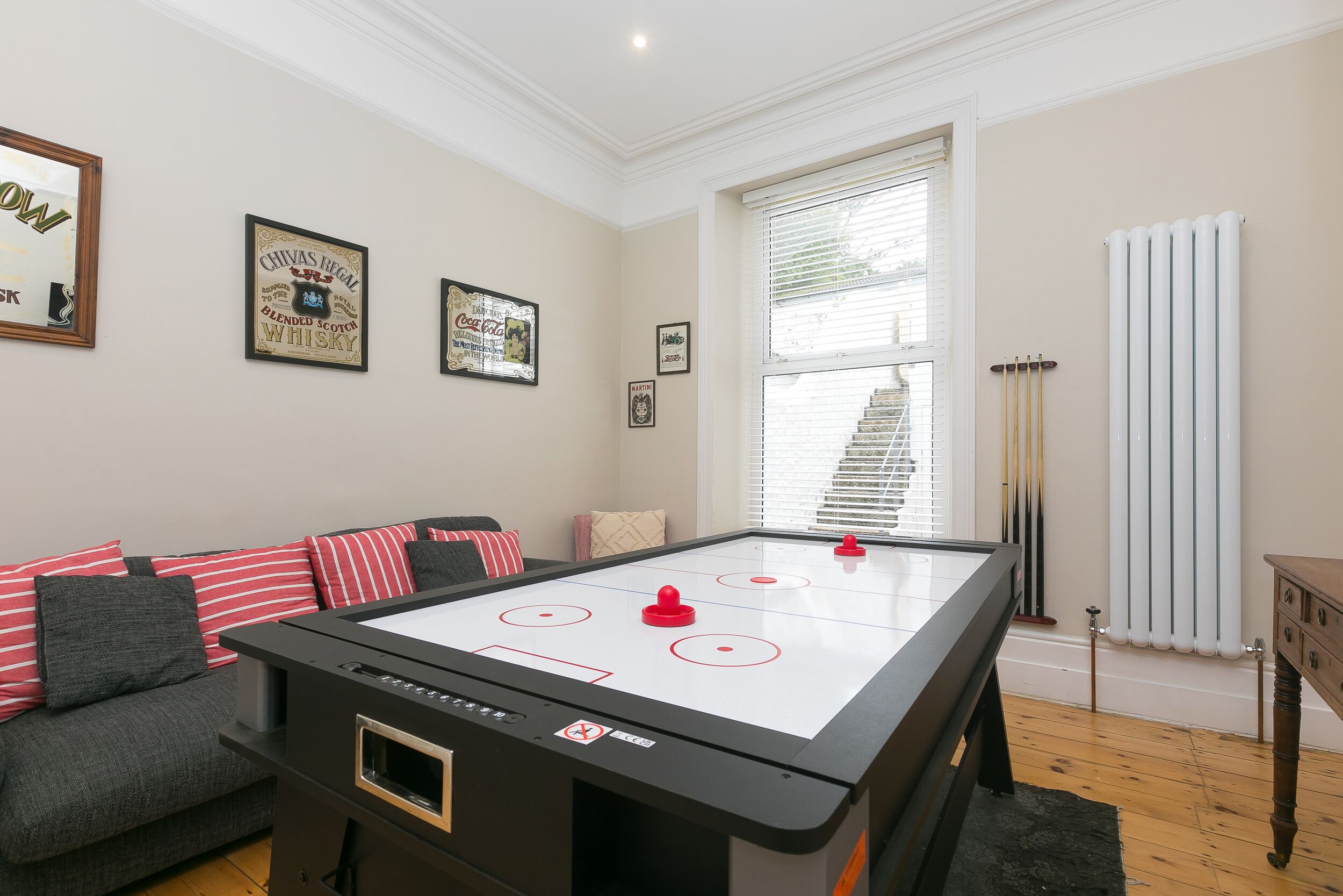 Ein Zimmer mit einem Billardtisch, einem grauen Sofa mit roten und schwarzen Kissen sowie gerahmten Bildern an der Wand.